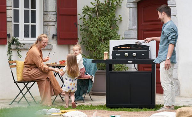 Famille cuisinant sur une plancha positionnée sur le chariot en acier avec tablettes Le Marquier